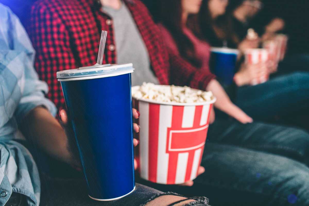 Close up of tasty but unhealthy food. There are basket of popcorn and a blue cup of coke on picture. Man and woman are holding it together.