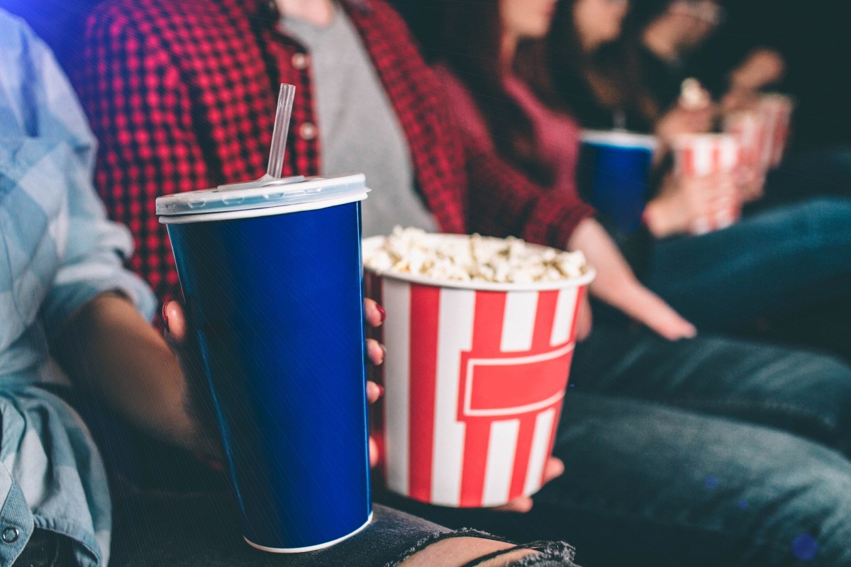 Close up of tasty but unhealthy food. There are basket of popcorn and a blue cup of coke on picture. Man and woman are holding it together.