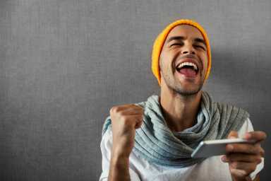 Happy handsome man with telephone. Win in a game. Gamer man wearing orange hat and gray scarf on gray background.
