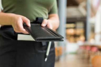 Cropped image of woman waitress in apron hold out bill folder to cafe visitor of cafe or restaurant.