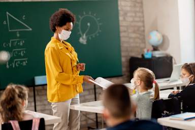 Happy black teacher giving exam paper to her student while wearing protective face mask in the classroom.