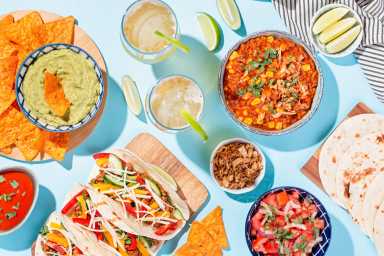 Mexican food table with traditional dishes. Chili con carne, tacos, tomato salsa, corn chips with guacamole. Mexican feast in hard light on blue color background, top view