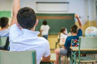 A boy and his classmates raise their hands as the teacher writes on the blackboard. Back to school concept