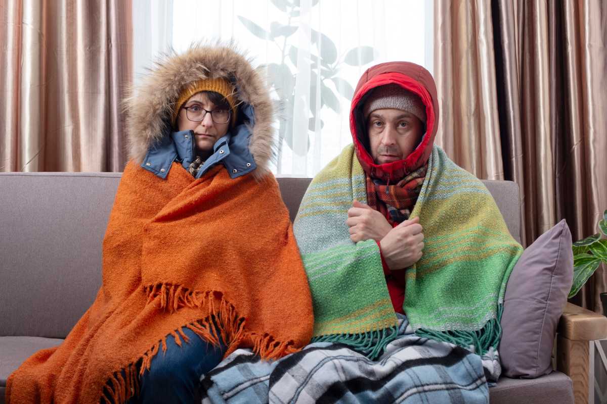 Sad middle age couple in warm clothes getting cold on the sofa in their living room