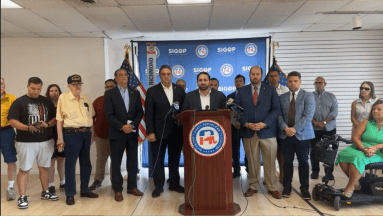 group of men and women inside a building at a podium speaking about attempted assassination of Donald Trump