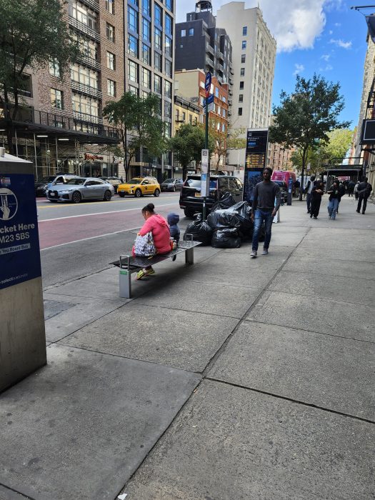 One of many NYC bus stops without shelter, outside during the day with two people sitting on it