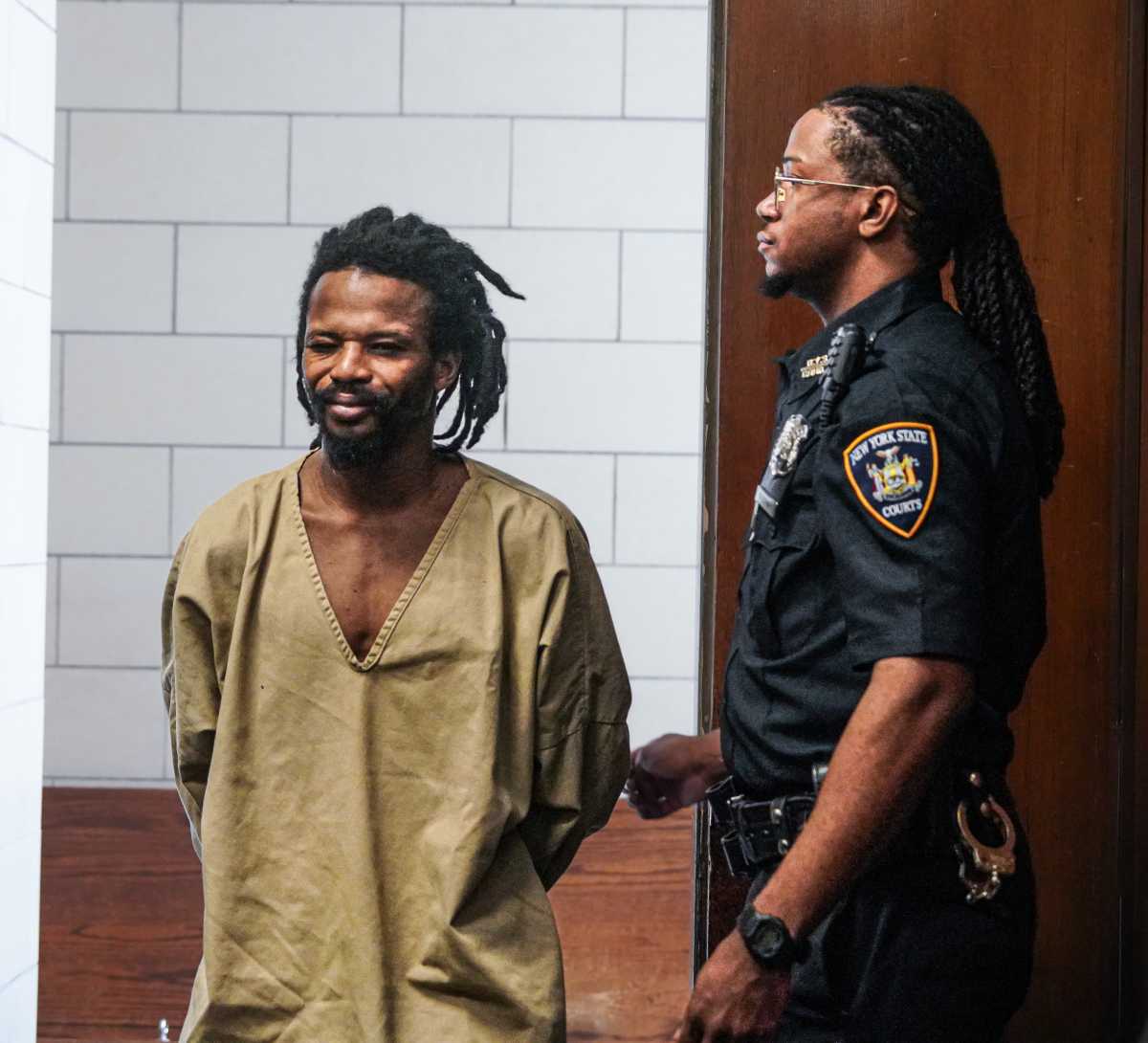 suspect in beige prison clothes with officer smirking during arraignment