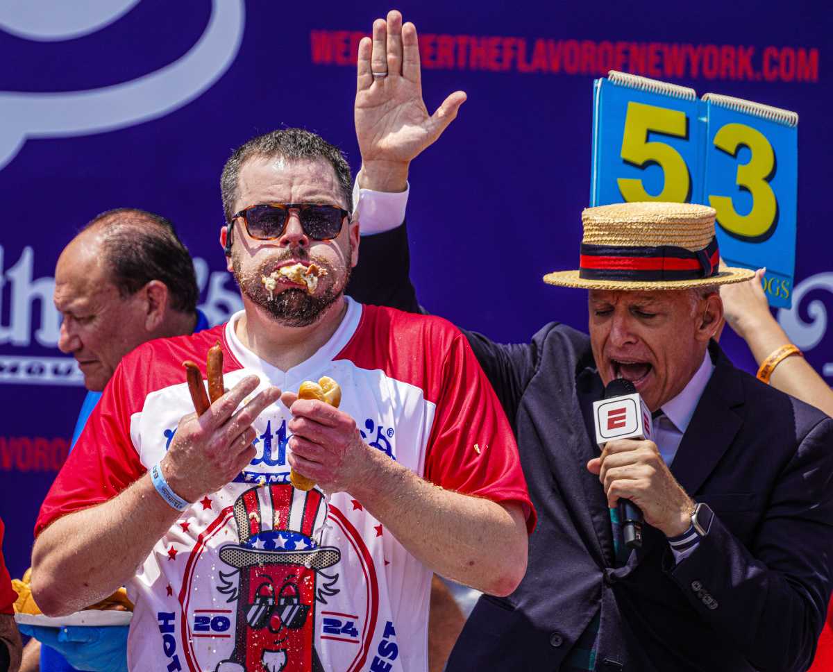 man eating hot dogs while man in microphone sings his praises