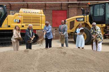 six people at a construction site holding shovels in front of dirt